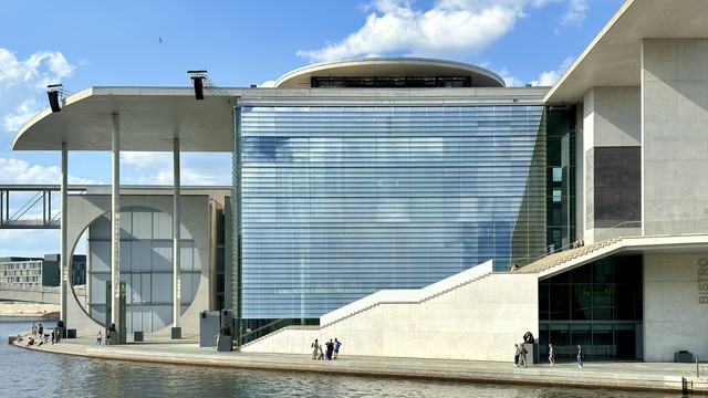 Modern building with large windows and geometric concrete structures alongside a river, with people walking and sitting nearby.