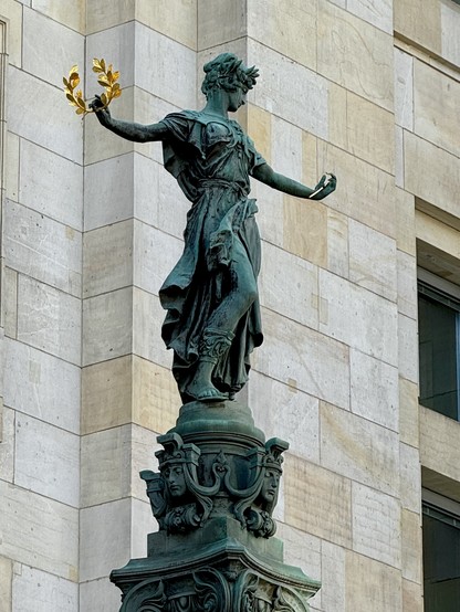 Statue of a robed figure holding a golden laurel wreath, mounted on a decorative pedestal in front of a stone building facade.