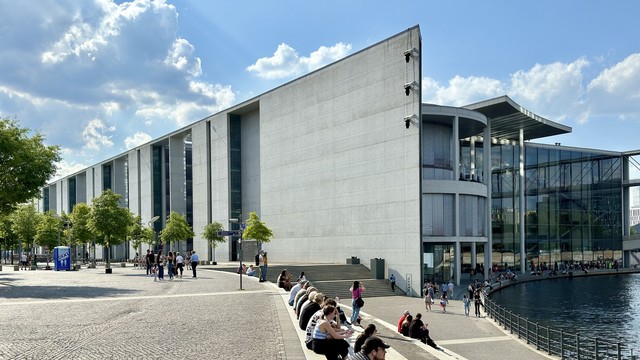 Modern building with large glass windows and a concrete facade. People are sitting on steps and walking around the area. There are trees and a body of water nearby. The sky is partly cloudy.