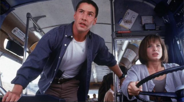 Screenshot from the movie "Speed", which was released on this day in 1994, showing Sandra Bullock sitting behind the steering wheel of a bus and Keanu Reeves standing next to her.