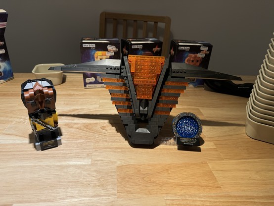 Lego-style models on a wooden table, including a spacecraft, a character figurine, and a circular emblem, with boxes and other objects in the background.