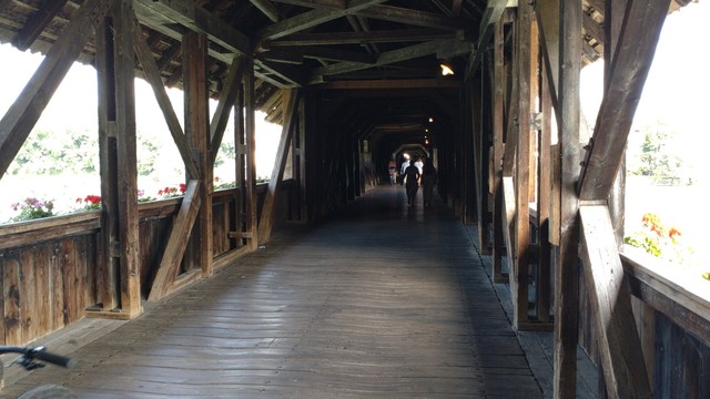 The wooden covered bridge over the Rhine between Bad Säckingen and Stein AG from the inside. Some side panels are open, some closed. Through the open ones you can see flower boxes with red flowers. Cyclists on the bridge in the distance.