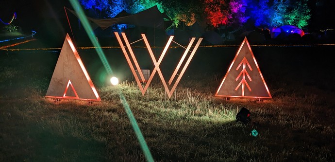 Das beleuchtete Vvoidcamp Logo aus Holz mit Zelten und RGB Beleuchtung auf Bäume im Hintergrund