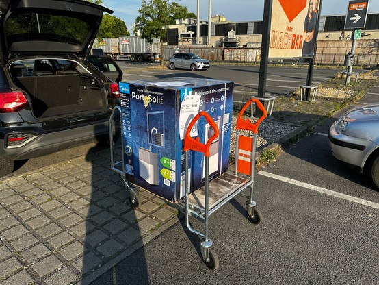 Ein riesengroßes Karton auf einem Einkaufswagen vor einem Auto