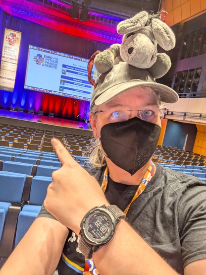 Me in the audience of Forum Hall at EuroPython, masked with my back to the front and my plush donkey on my head, pointing to the stage