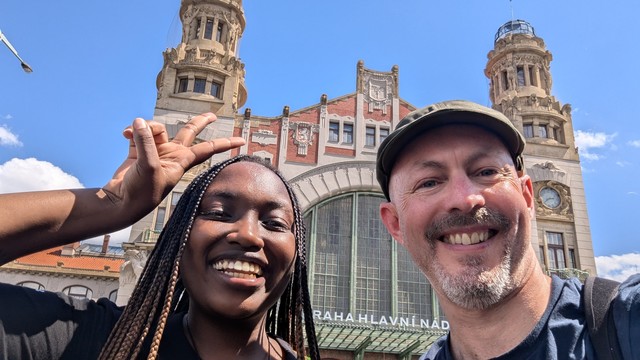 A photo of me and Velda standing in front of an impressive train station, with blue skies and fluffy white clouds in the background.