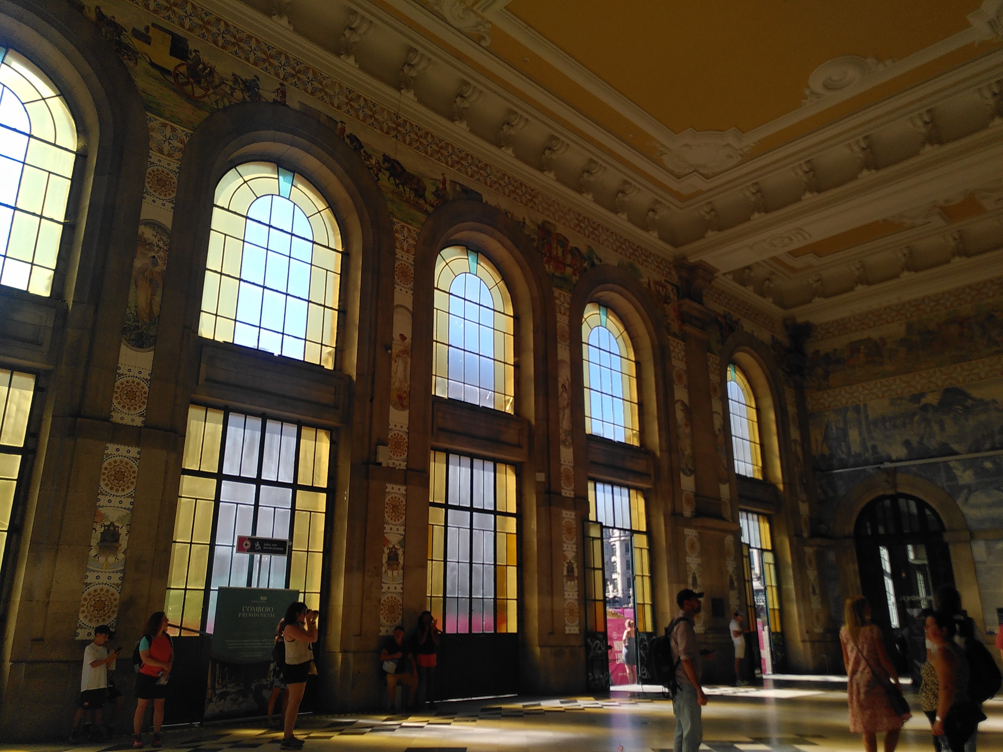 Abfahrtshalle. Die Wand nach außen. Hohe Fensterbögen bis unter die Decke. Dazwischen Fließen mit filigranen Ornamenten. Teilweise gelbes Fensterglas erzeugt eine andächtige Lichtstimmungen in der Halle.