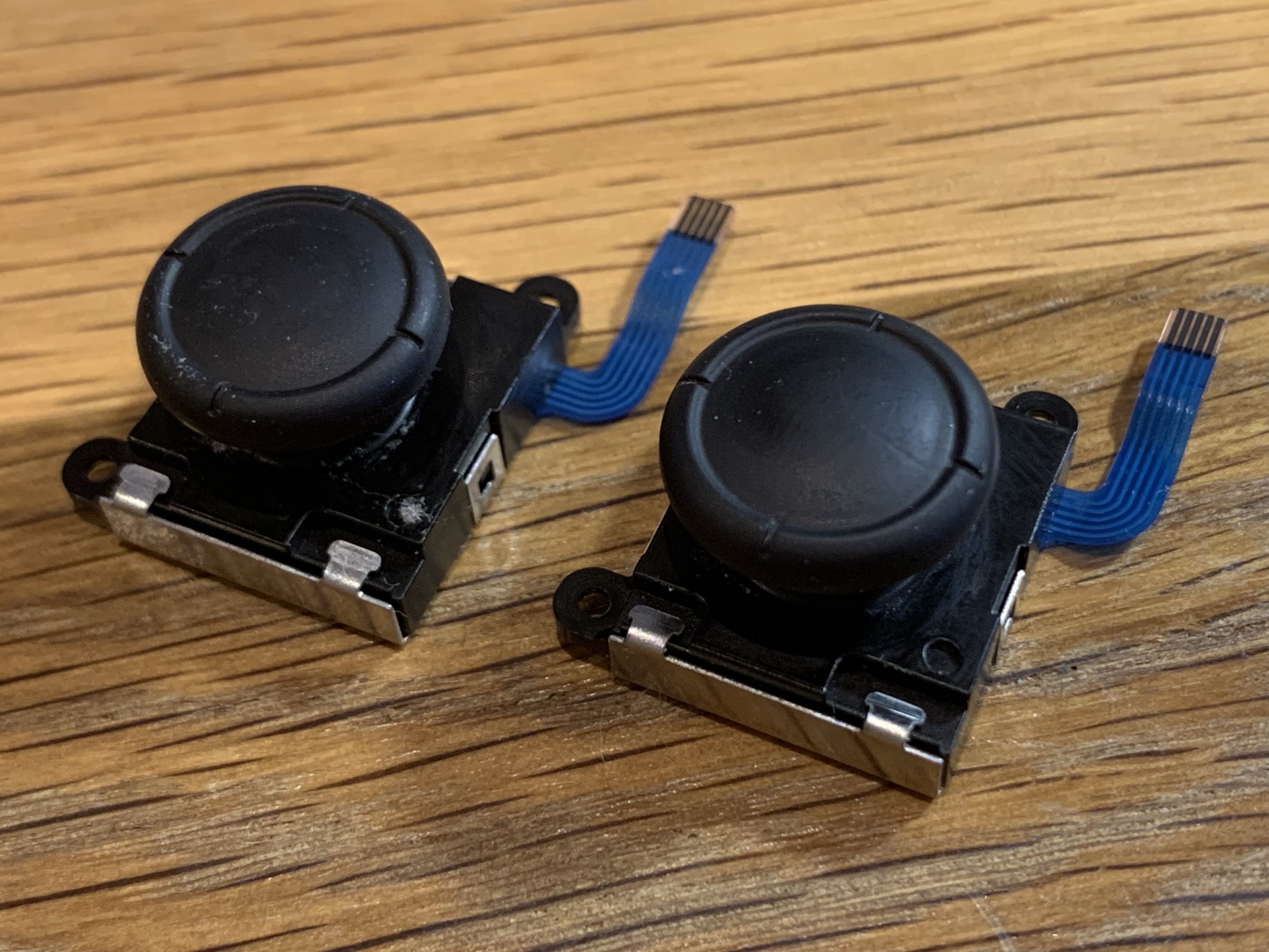 Two small joysticks with black buttons on top and blue wiring are displayed on a wooden surface.