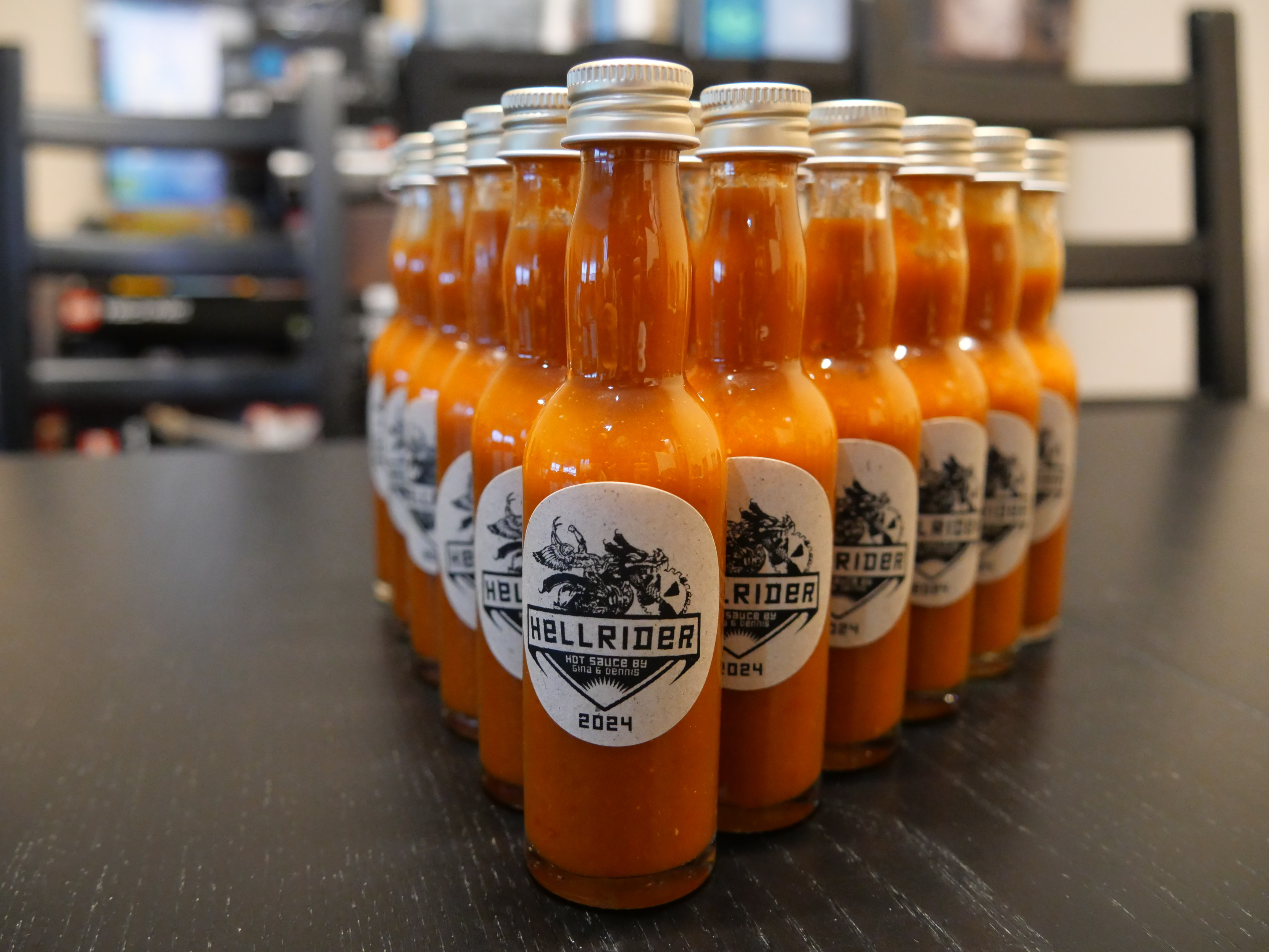 A bunch of small hot sauce bottles on a table. The bottles are labelled. The labels say 
    
    Hellrider
    Hot sauce by Gina & Dennis
    2024