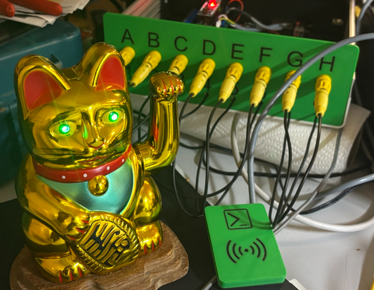 A Maneki-neko with green glowing LEDs as eyes. In the Background a strange Device, maybe part of this years challenge.