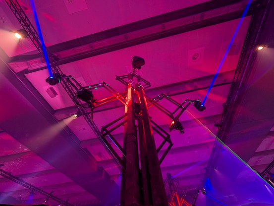 Looking up towards one of the big metal towers full of lamps. Its rusty metal, pink lid shining blue beams of lights like eyes.