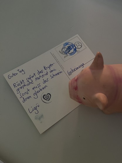 Ein Plastikschwein steht neben einer Postkarte, auf der steht: „Guten Tag. Rückt sofort das Kryptographische Material raus, sonst muss das Schwein dran glauben. Ligrü“