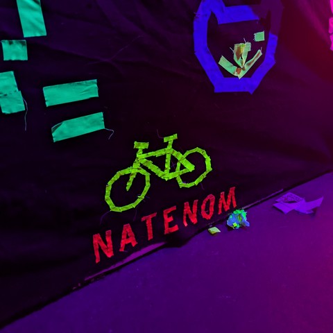 Sinnbild Fahrrad mit dem Schriftzug Natenom aus fluoreszierendem Klebeband an einer dunklen Wand.