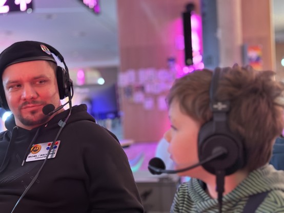 Ein Mann in einem schwarzen Hoodie und einer Baskenmütze trägt ein Headset mit Mikrofon, während er einen kleinen Jungen ansieht, der auch ein Headset aufgesetzt hat. Der Hintergrund zeigt bunte Lichter.