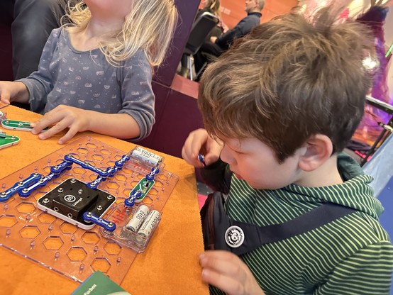 Zwei Kinder interagieren mit einem pädagogischen Schaltungsbausatz auf einem Tisch. Das Kit enthält farbenfrohe Komponenten, die auf einem klaren, sechseckig gemusterten Brett angeordnet sind. Der Hintergrund zeigt andere Personen, die an Aktivitäten beteiligt sind.