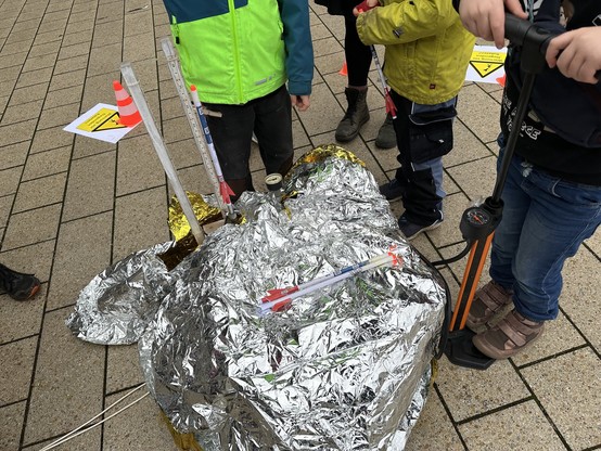 Eine Gruppe von Kindern steht neben einem mit Silberfolie bedeckten Objekt, möglicherweise einer Rakete, mit Messstäben und einer Pumpe. Auf dem Boden sind leuchtend orangefarbene Verkehrskegel und Warnschilder zu sehen.