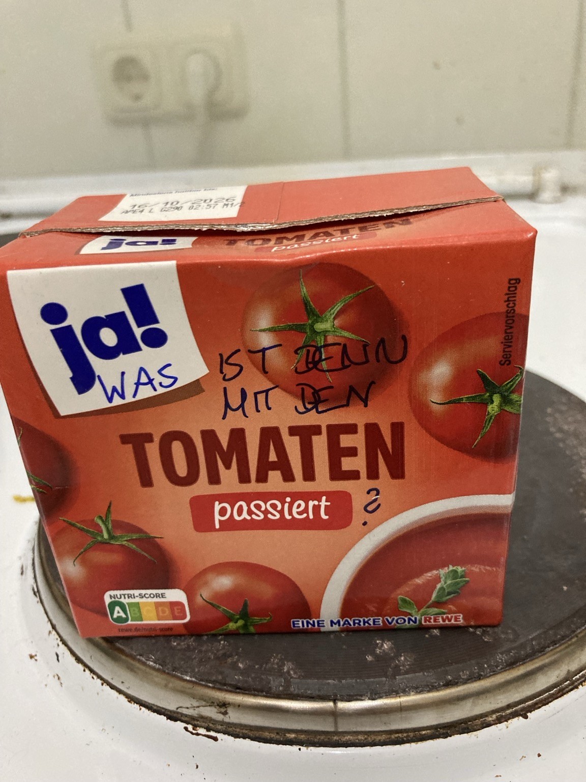 A red carton labeled "passiert Tomaten" (strained tomatoes) with a handwritten note asking "Was ist denn mit den?" (What is up with them?). The carton is placed on a stovetop with a visible burner.