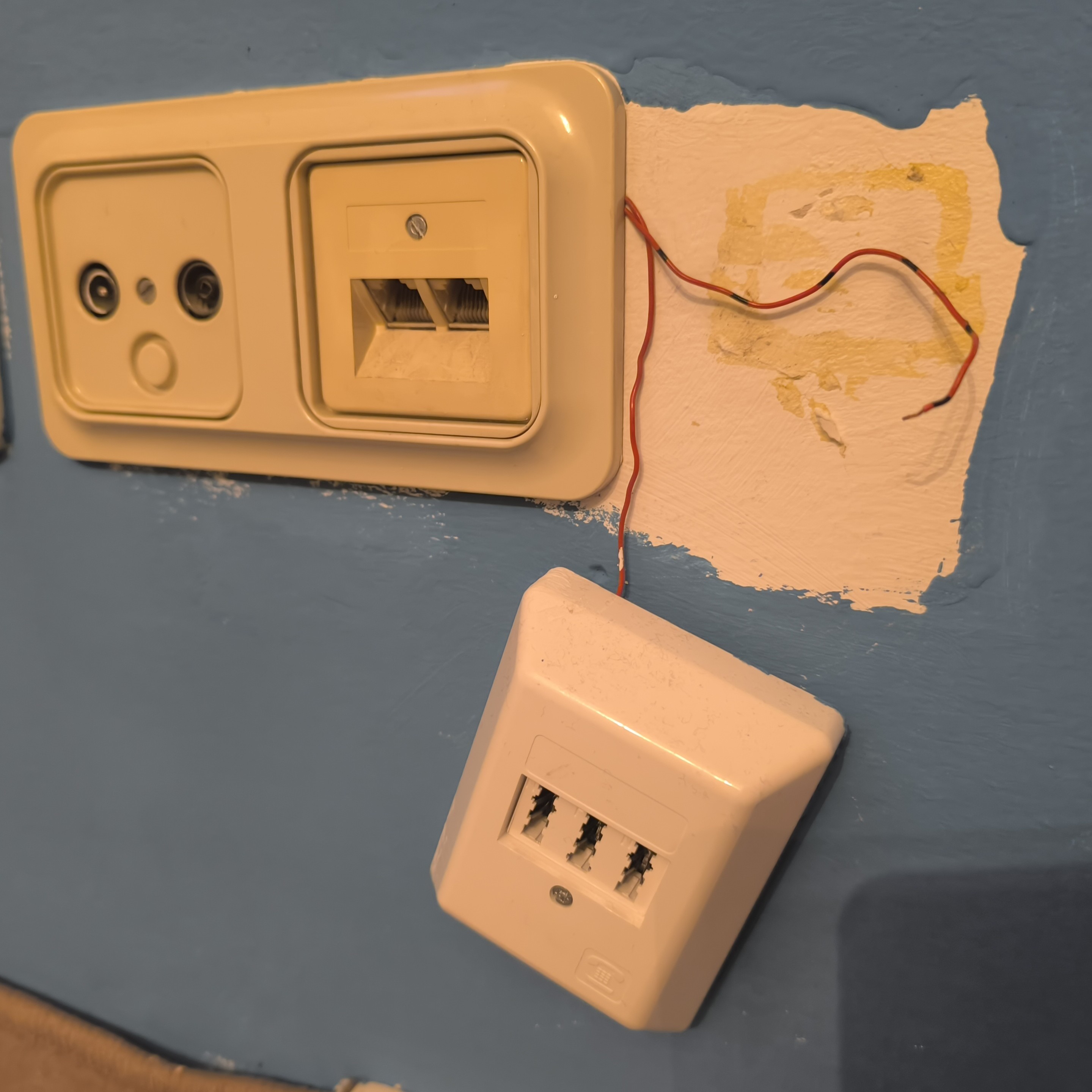 Foto einer Wand, von der eine TAE-Dose halb herabgefallen ist. Sie war offenbar nur mit einem Power Strip befestigt. Das Kabel baumelt lose herum und wurde einfach hinter der Blende aus einer daneben ordentlich montierten Dose rausgeführt.