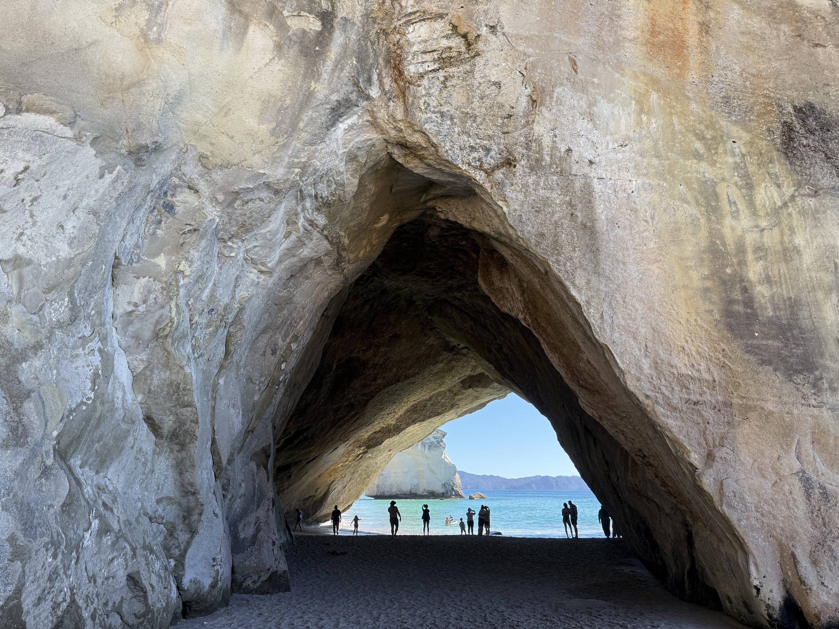 A natural rock archway leading to a beach, with several silhouettes of people walking along the shore and ocean visible in the background. The scene features sandy ground and cliffs with textured rock formations. A natural rock archway leading to a beach, with several silhouettes of people walking along the shore and ocean visible in the background. The scene features sandy ground and cliffs with textured rock formations.
