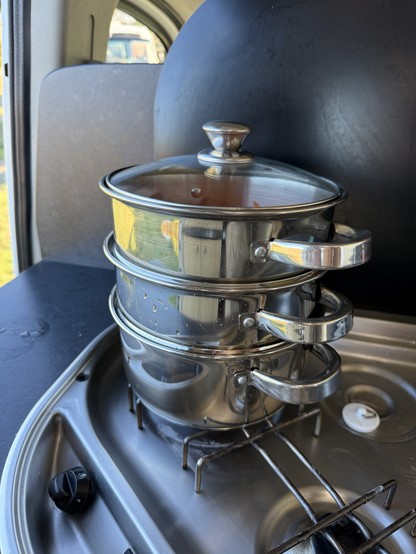 Three stacked stainless steel pots are on a gas stove. The top pot has a lid, and the stovetop features a knob and a circular burner. The scene is set in a van or camper.