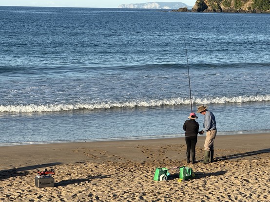 A person and an adult are fishing on a sandy beach, facing the ocean. The adult is assisting the person with their fishing rod by using a cordless screwdriver. Nearby, there are two bags on the sand and a green box with fishing equipment and a drone. The background features calm waves.