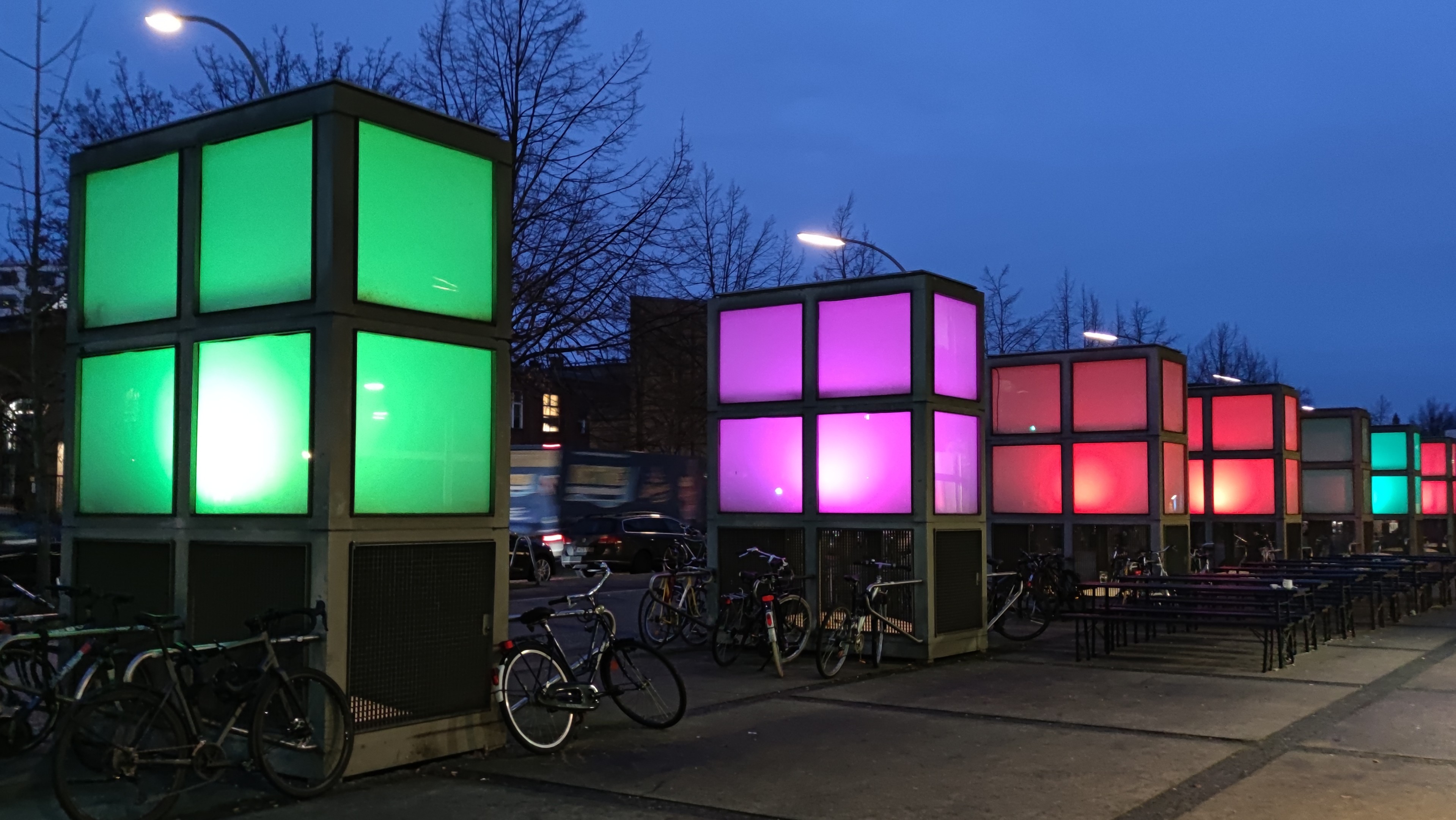 Quaderförmige Leuchtelemente mit quadratischen Scheiben stehen entlang eines Fußwegs. Quaderförmige Leuchtelemente mit quadratischen Scheiben stehen entlang eines Fußwegs.