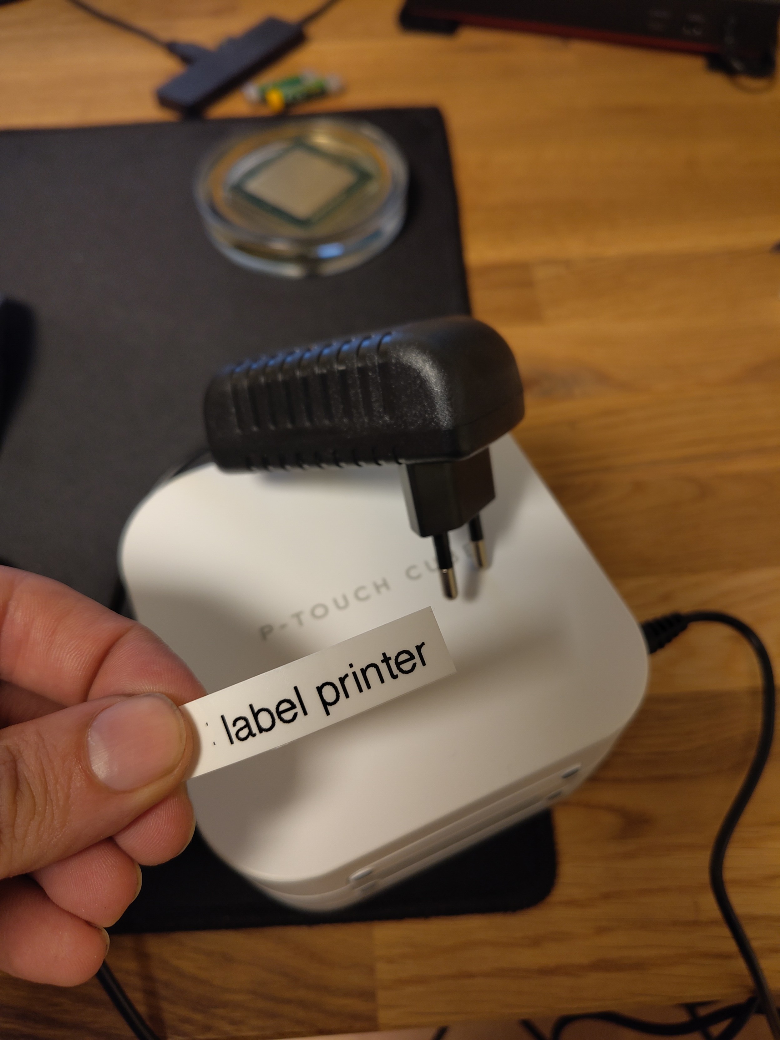 A brother p-touch cube label printer with a power supply on my desk, I hold a label with "label printer" printed on it in my hand