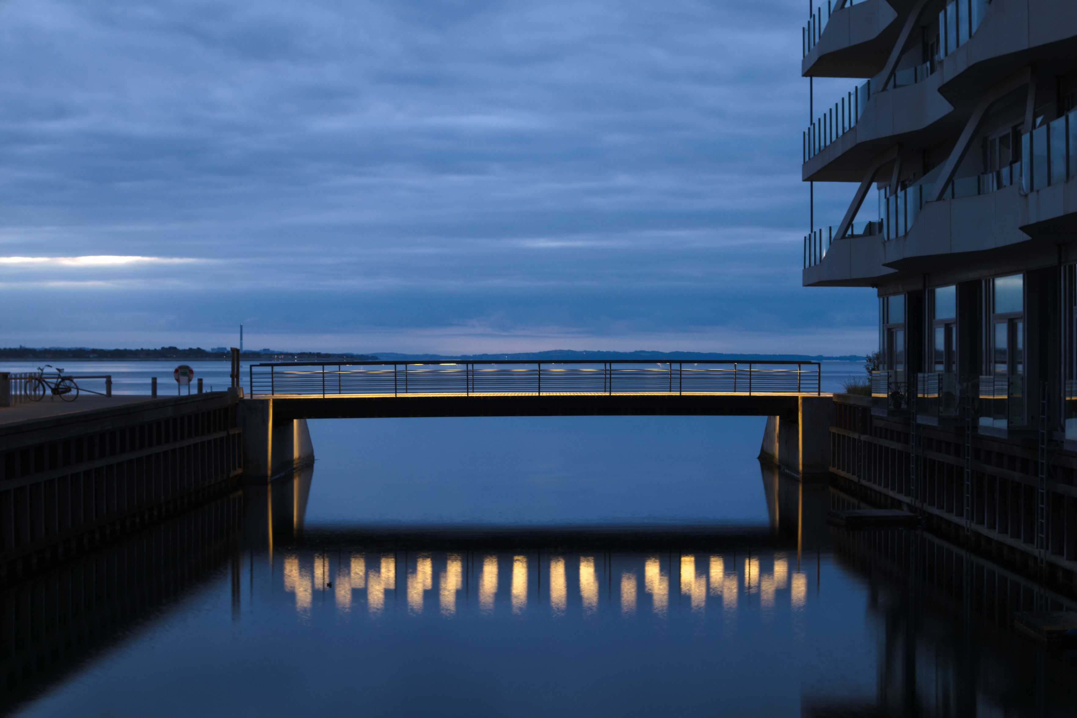 Eine beleuchtete Brücke über einem Kanal mit dem Horizont im Hintergrund. Der Baustil ist modern und kantig. Am rechten Bildrand ist ein Wohngebäude zu sehen. Die orange Beleuchtung der Brücke spiegelt sich in dem ruhigen Wasser und setzt einen farblichen Kontrast zu den Blautönen, die das restliche Bild beherrschen. Eine beleuchtete Brücke über einem Kanal mit dem Horizont im Hintergrund. Der Baustil ist modern und kantig. Am rechten Bildrand ist ein Wohngebäude zu sehen. Die orange Beleuchtung der Brücke spiegelt sich in dem ruhigen Wasser und setzt einen farblichen Kontrast zu den Blautönen, die das restliche Bild beherrschen.