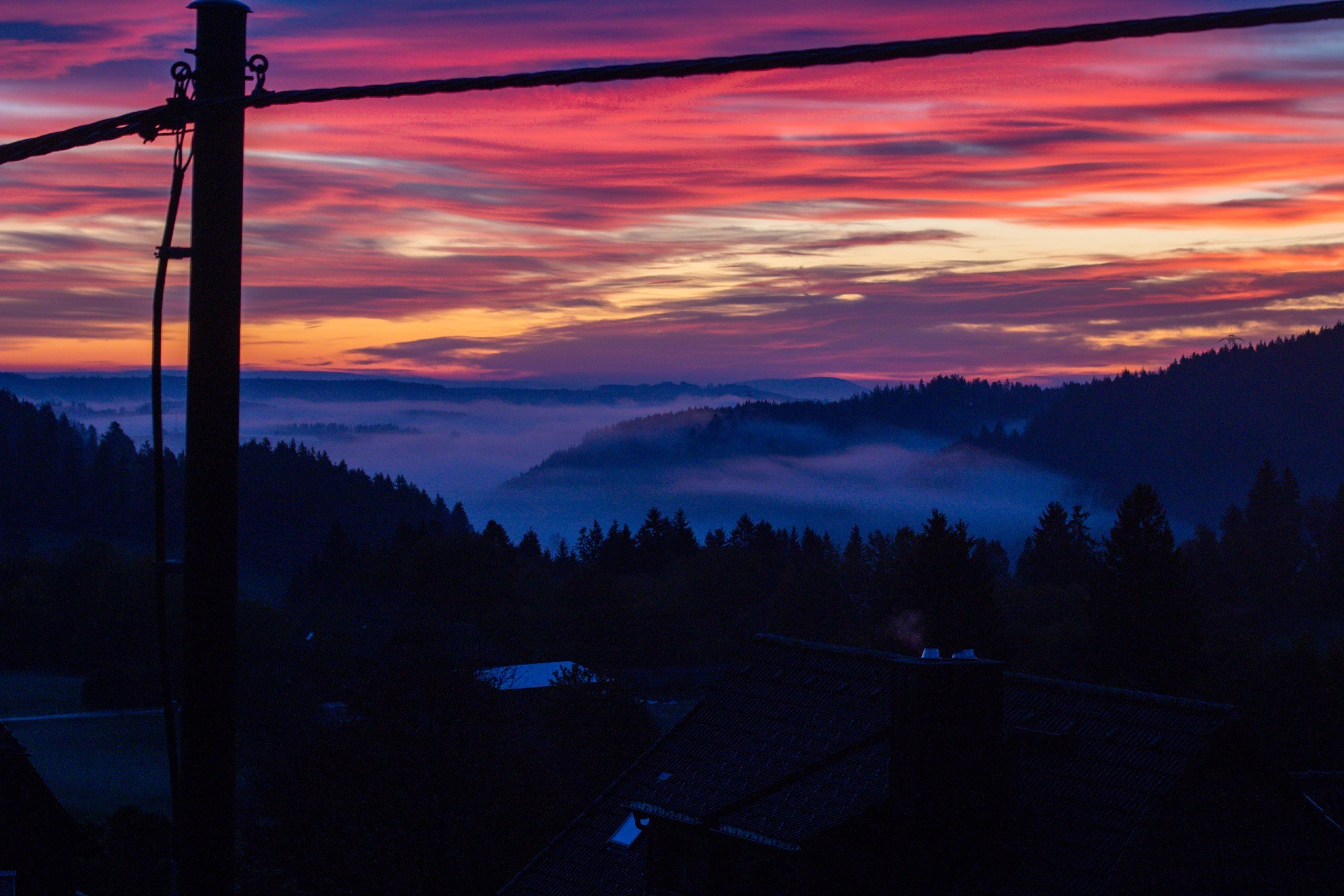 A vibrant sunset casts pink and orange hues across the sky, with layers of fog blanketing the distant hills and valleys. In the foreground, silhouettes of trees and rooftops are visible, along with a power pole and power lines framing the scene. A vibrant sunset casts pink and orange hues across the sky, with layers of fog blanketing the distant hills and valleys. In the foreground, silhouettes of trees and rooftops are visible, along with a power pole and power lines framing the scene.
