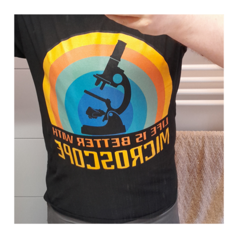 Foto, Tshirt an meinem Körper, schwarz, unten ein schriftzug "Life is better with microscope", darüber eine schearze siluette eines älteren Mikroskop mit geradem tubus, angekipptem tisch und Spiegel. Dahinter sind konzentrische kreise aus Farebn, außen beginnend mit gelb und dann nach innen fortsetzend über orange türkis, petrol bis hin zu blau in der mitte.