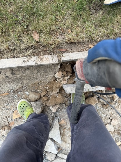 Eine Person steht auf unebenem Boden in grünen Crocks und dunklen Hosen und hält ein Elektrowerkzeug über einem teilweise zerbrochenen Betonabschnitt. Schmutz und Felsschutt sind verstreut, und Gras ist im Hintergrund sichtbar.