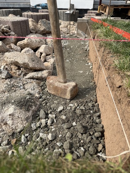 Eine Baustelle mit einem Holzhammer, der auf einem Bett aus Kies und Schmutz ruht. Zu den umliegenden Materialien gehören große Felsen und Betonblöcke, mit einer roten Schnur, die die Standortgrenze markiert, und einem Stück Gras im Vordergrund.