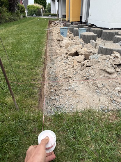 Eine Baustelle, die einen Graben zeigt, der durch eine Schnurlinie zur Ausrichtung markiert ist. Auf der einen Seite gibt es Gras, während die andere Seite eine Mischung aus Kies und Schutt hat, mit mehreren Eimern und Betonblöcken sichtbar.