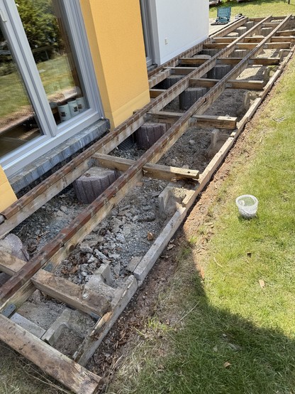 Baustelle mit freiliegendem Holzrahmen neben einem Haus. Das Gebiet verfügt über eine Mischung aus Kies und Schutt, was auf laufende Renovierungs- oder Fundamentarbeiten hinweist. Gras ist neben der Bauzone sichtbar.