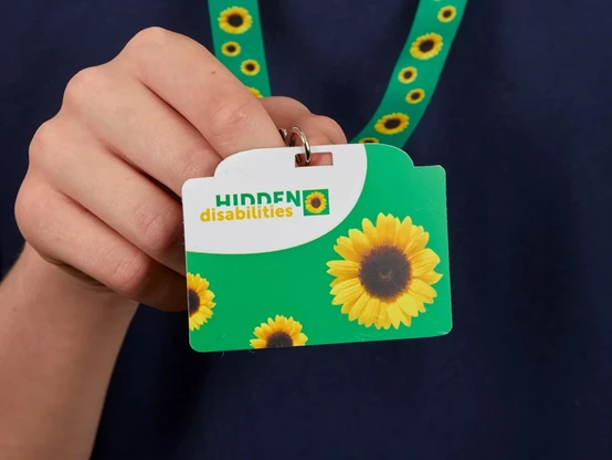 Nahaufnahme eines Lanyards mit Kreditkartenformat-Anhänger. Der Bändel ist grün und mit Sonnenblumen bedruckt. Auf dem Anhänger sind ebenfalls Sonnenblumen auf grünem Hintergrund abgebildet, und der Text hidden disabilities