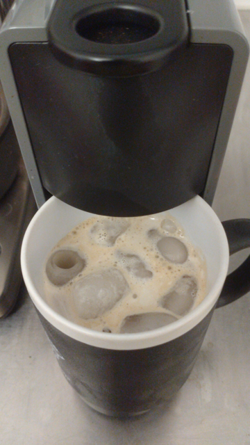 A black mug with white inside (yes, it's an Easterhegg 2019 Vienna mug) filled with (round) ice cubes and coffee with still some crema on top standing under the outlet of a narrow Nespresso capsule coffee machine.