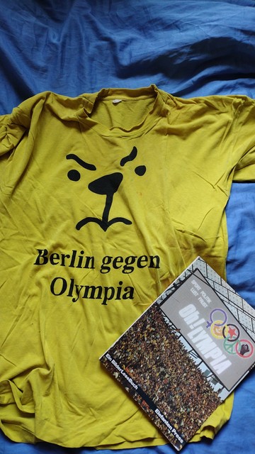 Gelbes T-Shirt der Kampagne gegen die Bewerbung Berlins für Olympia 2000 mit der Strichzeichnung eines grumpeligen Bärengesichts, darunter der Schriftzug "Berlin gegen Olympia". Das T-Shirt hat bessere Tage gesehen. Diverse Flecken zeugen davon, dass es zwischendurch als Renovierungsshirt herhalten musste. Das liegt das Buch "Oh!lympia", das im Rahmen der damaligen Kampagne bei Elefanten Press erschien.