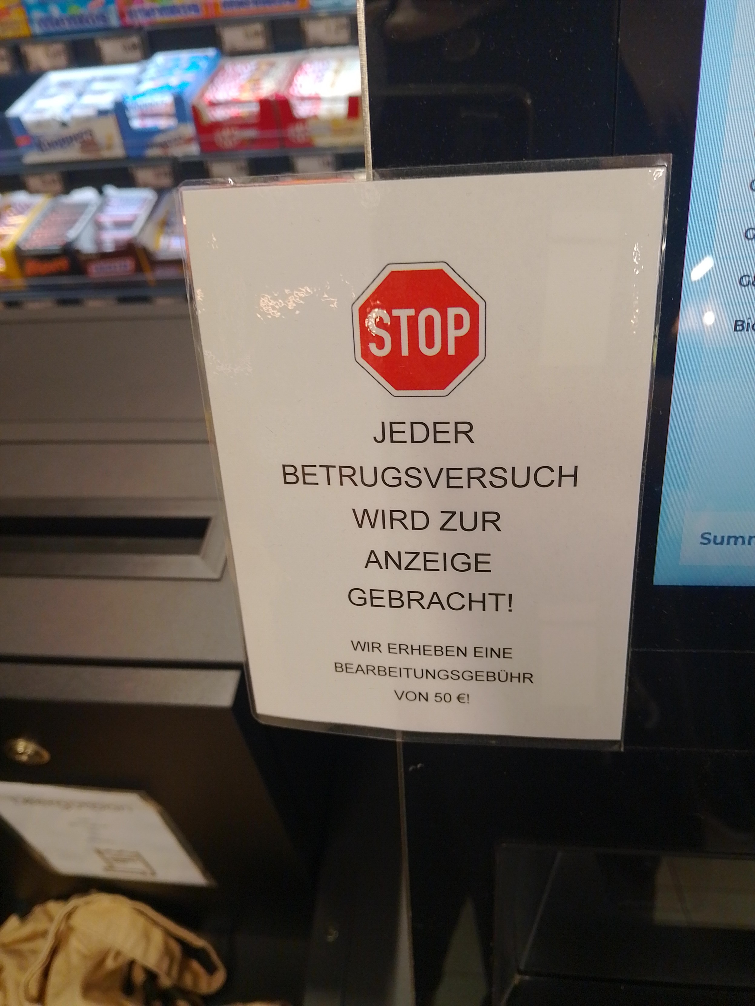 Schild mit einem Stopschild darauf. Darunter steht: Jeder Betrugsversuch wird zur Anzeige gebracht. Dieses Schild klebt an einer SB Kasse