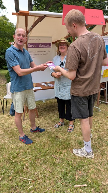 Drei Personen stehen im Freien an einem Infostand mit dem Banner „Save Social – Soziale Netzwerke als demokratische Kraft stärken“. Eine Frau in der Mitte trägt einen Sonnenhut und lächelt, während zwei Männer Flyer austauschen. Im Hintergrund ist ein weiteres Schild mit der Aufschrift „Tactical Tech“ zu sehen. Der Stand befindet sich auf einer Wiese, es ist sommerlich und freundlich. Der Fokus liegt auf dem Austausch von Informationen in einer offenen, einladenden Atmosphäre.

