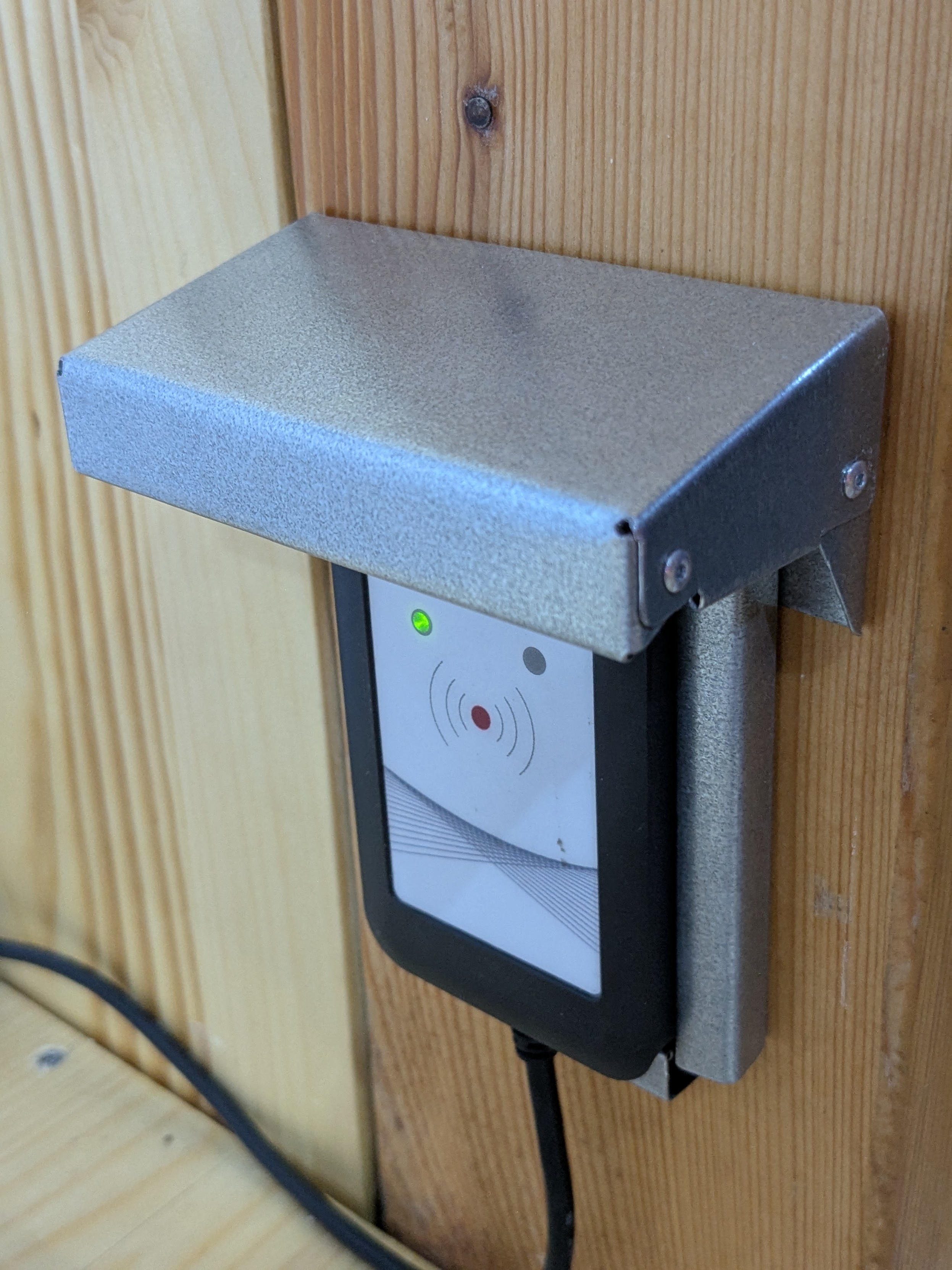 A picture of an RFID reader mounted to a wooden beam. There is a roof over the reader to protect it from water leaking from above. The roof was bent from sheet metal and was assembled using blind rivets.