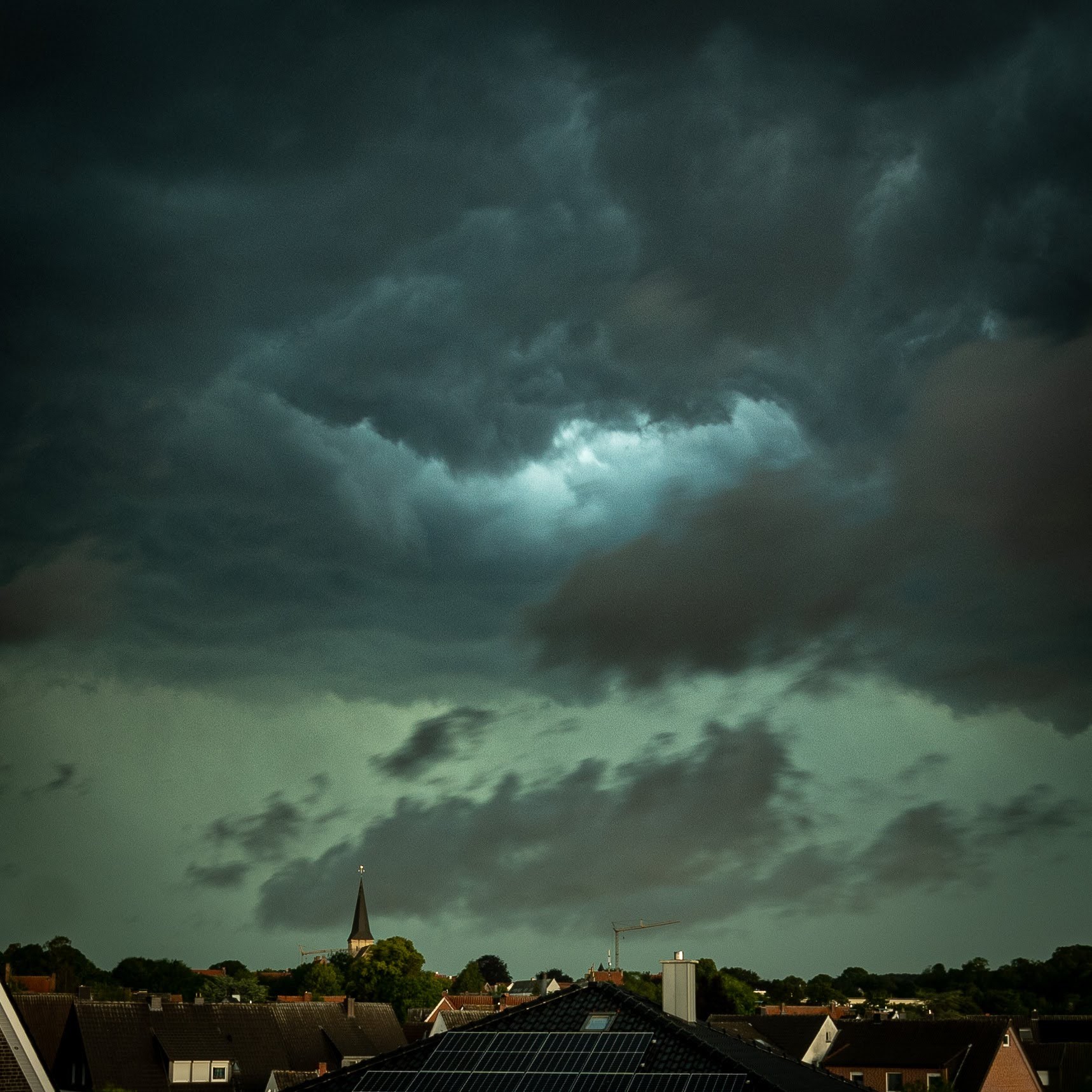 Über den Dächern einer Dorfes im Münsterland naht eine grün blaue Wolke mit einem hellen Zentrum heran.
