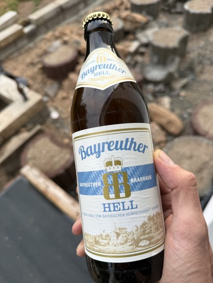 Eine Person hält eine Flasche Bayreuther Hell in der Hand. Das Etikett zeigt blaue und goldene Farben mit dem Text "Bayreuther Hell" und einer Illustration einer Landschaft. Der Hintergrund zeigt verschwommene Holzstümpfe und Bauschutt.