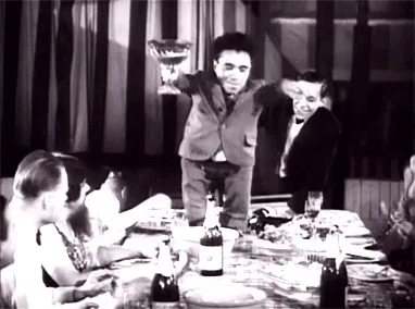 Scene from black and white movie, one person dancing on a table, holding a cup. Everyone chanting "Gooble Gobble One of Us"