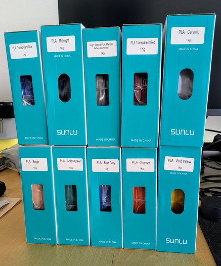 Ten boxes of 1kg PLA filament roles in different colors bought in a sale stacked on a desk
