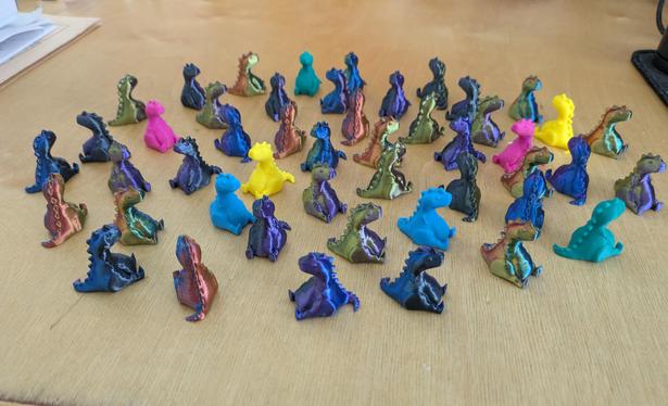 A large number of tiny 3d printed chubby T-Rex models grouped together on a table showing off the different filament colors.