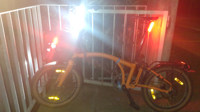 Mein orangenes Brompton Electric G-Line an ein eiserenes Geländer gelehnt in Vollbeleuchtung inklusive roten Knog Plus Leuchten seitlich an die Seitentaschen der Fronttasche geklipst.