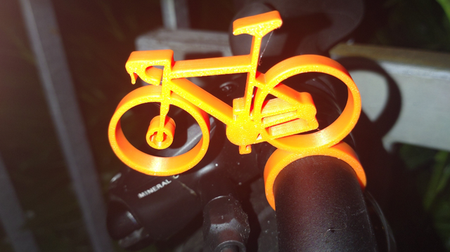 Ein stehendes, vowärts gerichtetes, orangefarbenes 3D-gedrucktes Rennrad am schwarzen Lenker des Brompton G-Line festgeklipst.
