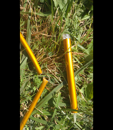 Ein gebrochenes, goldgelbes Zeltstangenteilstück im Gras, daneben das für diesen Bruch zu enge Reparaturstück.