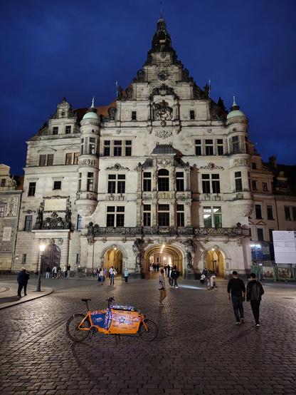 Das orange Lastenrad steht mit Lichterkette geschmückt auf dem beleuchteten Schlossplatz vor dem Georgentor in Dresden.