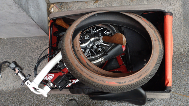 Blick von oben in einen Fahrradanhänger. Darin liegt ein zusammengefaltetes, weiss-rotes 16" Brompton und zwei 20 Reifen mit kleinen Noppen und brauner Seitenwand.